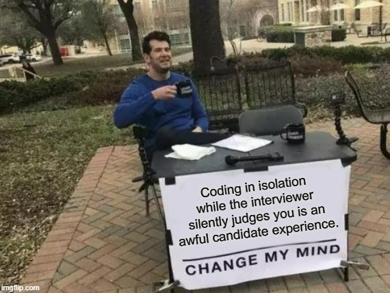 Un homme assis derrière un bureau avec un panneau indiquant "changez d'avis : coder en isolation pendant que l'interviewer vous juge en silence est une expérience terrible pour un candidat".